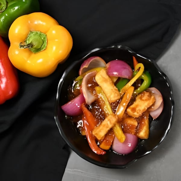 Chaska's chili paneer with bell peppers and onions in a dark bowl, with whole peppers nearby.
