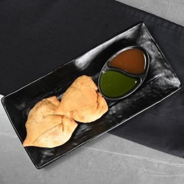 Two golden-brown Chaska samosas on a black rectangular plate with green and brown chutneys.
